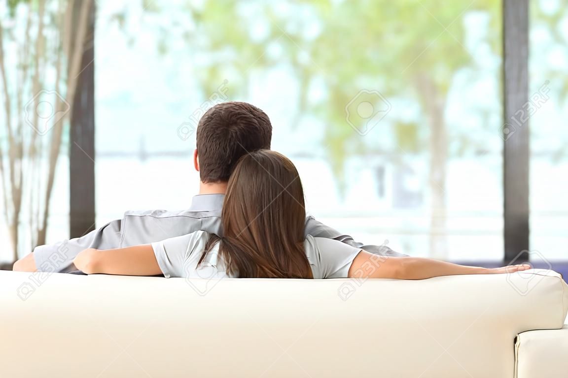 ソファに座って、屋外リビング ルームの窓からの緑の背景を探しているカップルを抱き締めることの後姿
