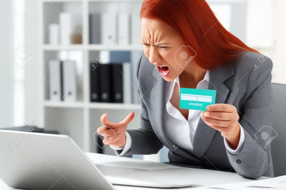 Angry executive woman pays online with credit card on laptop sitting on a desk at the office
