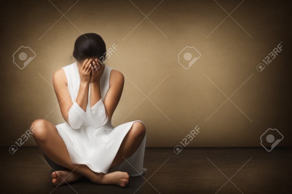 Vrouw zit op een oude vloer met scheuren. Ze is verdrietig en depressief, bedekt haar gezicht met handen. Studio papier achtergrond achter haar.