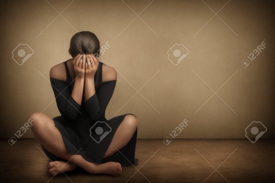 Vrouw zit op een oude vloer met scheuren. Ze is verdrietig en depressief, bedekt haar gezicht met handen. Studio papier achtergrond achter haar.