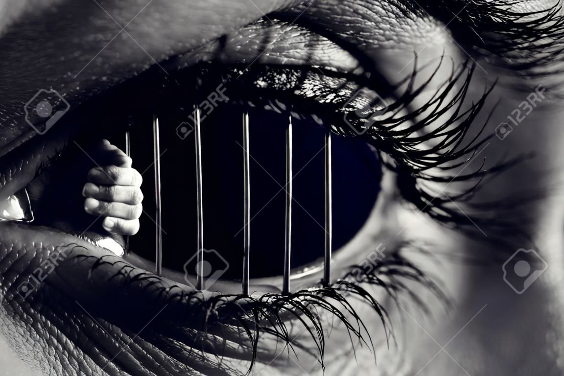 Conceptual monochrome photo of hands holding the bars of a prison inside a human eye