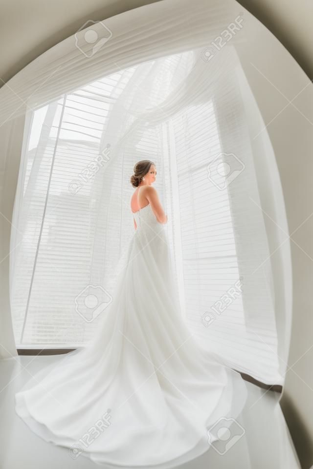 Gyönyörű fiatal menyasszony a fehér menyasszonyi ruha beltérben. Luxuty modell álló teljes hosszúságú ellen nagy ablak otthon. Lány vár a vőlegénye, és nézi kamera világos szobában. Magas kulcs, készült halszem objektív fotó.