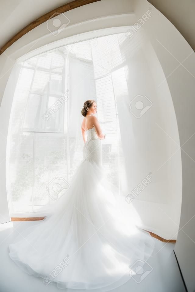 Gyönyörű fiatal menyasszony a fehér menyasszonyi ruha beltérben. Luxuty modell álló teljes hosszúságú ellen nagy ablak otthon. Lány vár a vőlegénye, és nézi kamera világos szobában. Magas kulcs, készült halszem objektív fotó.