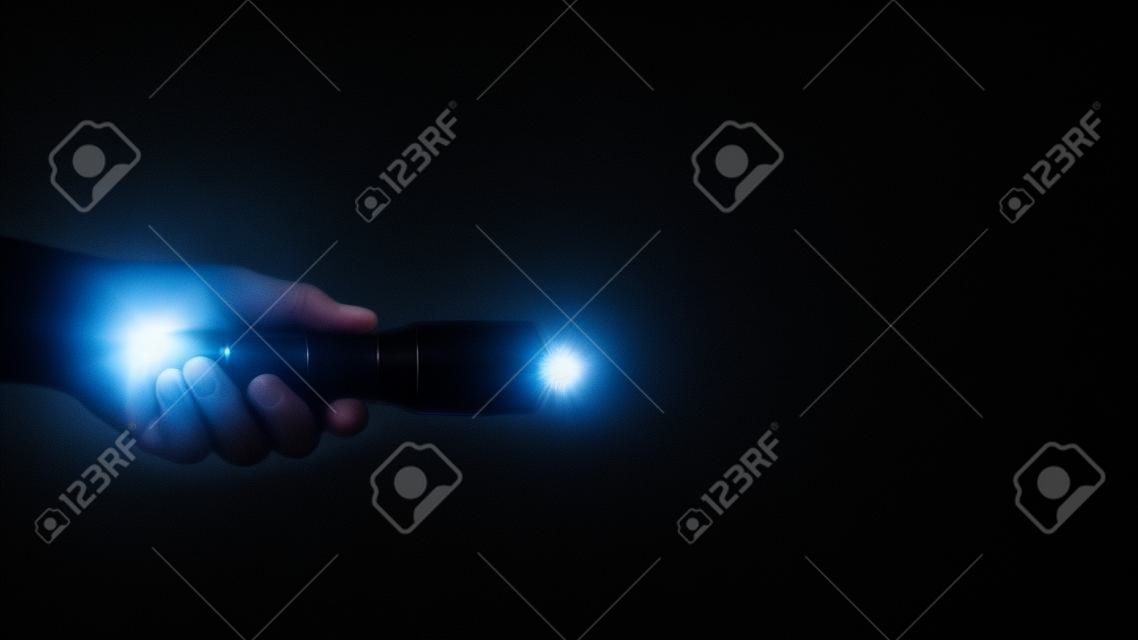 Schwarze Taschenlampe in Menschenhand auf schwarzem Hintergrund, darunter ein weißer Strahl.