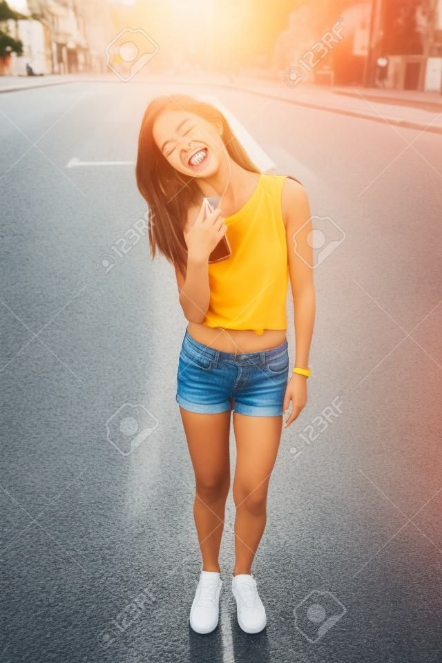 Ragazza gioiosa che balla per strada in una giornata di sole tenendo le mani in alto con smartphone e auricolari