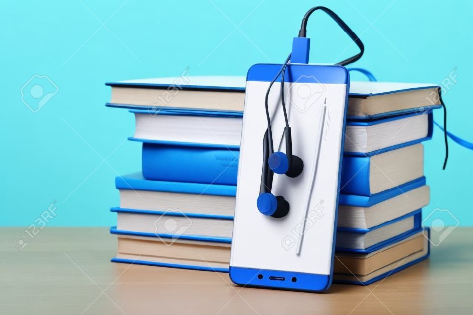 Telefono con auricolari bianchi accanto a una pila di libri su sfondo blu. Il concetto di audiolibri e educazione moderna
