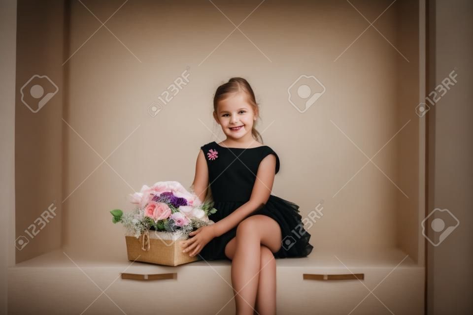 검은 드레스를 입은 작고 귀여운 소녀가 꽃다발을 들고 앉아 웃고 있습니다.