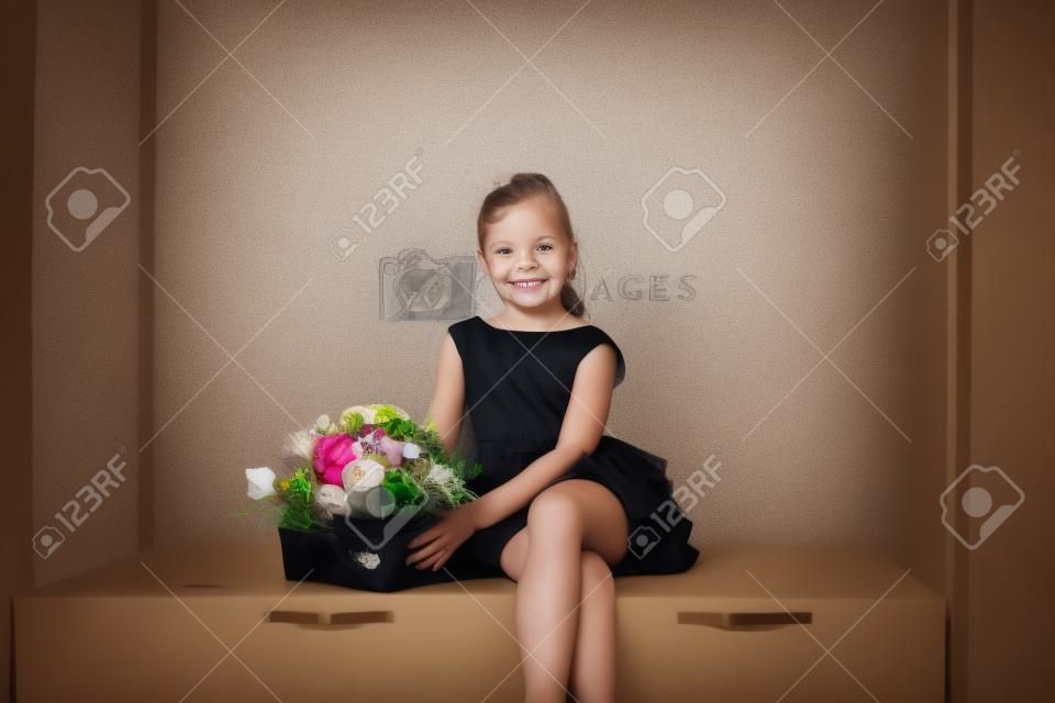 A little cute girl in a black dress is sitting and smiling with a bouquet of flowers.