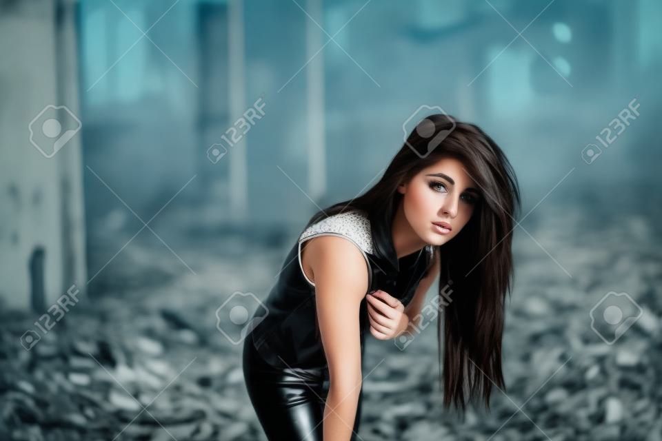 Portrait of young cute brunette girl wearing on black leather pants and white blouse posed on abandoned place.