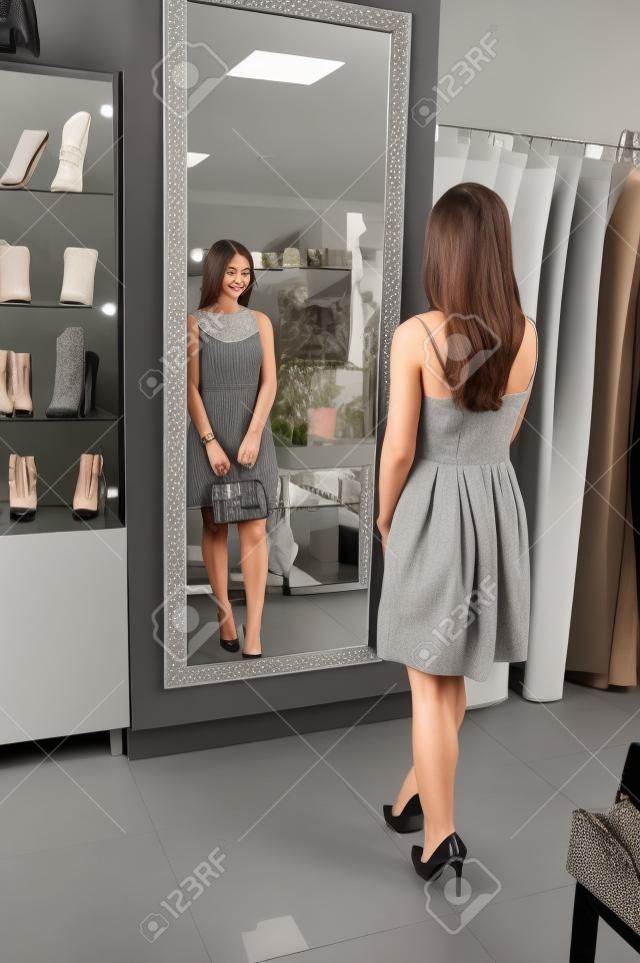 Portrait of a fantastic young woman in grey dress trying on new shoes and handbag in front of a mirror in shop.