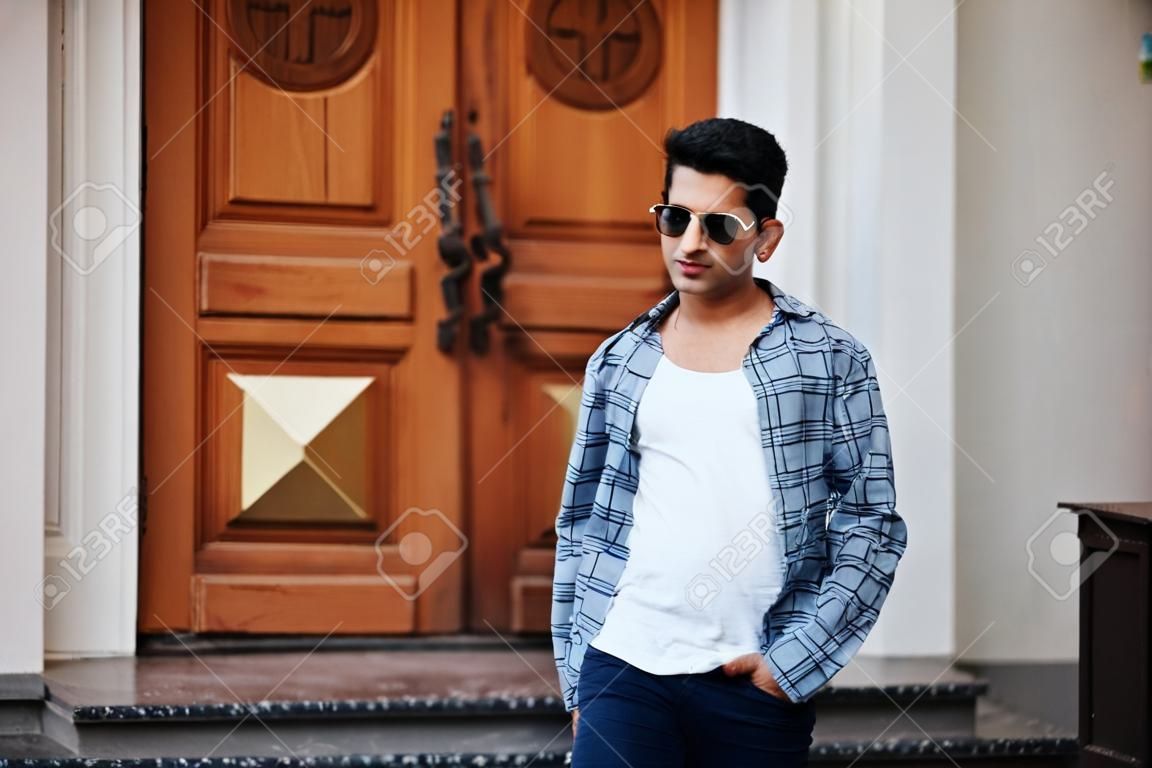 Stylish indian young man at sunglasses wear casual posed outdoor against door of building, show finger at camera.