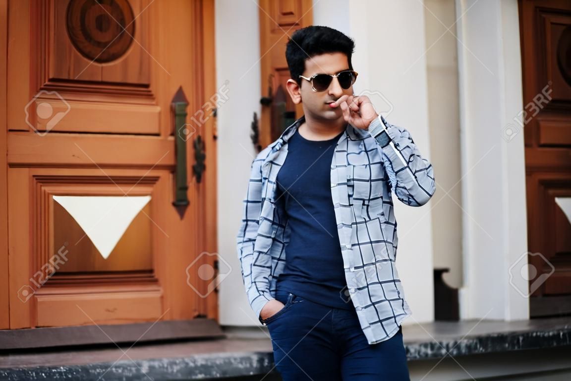 Stylish indian young man at sunglasses wear casual posed outdoor against door of building, show finger at camera.