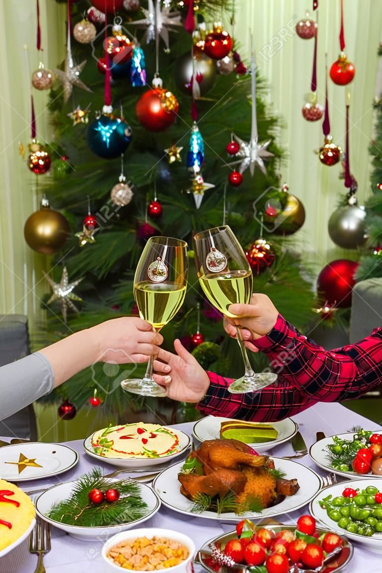 Human hands holding glasses with sparkling wine against beautiful decorated Christmas tree and served table with tasty dishes. New Year wishes with champagne. Couple celebrate Christmas at home.