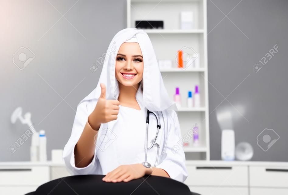 Consulta em clínica de cosmetologia. Médico de beleza feminino em casaco branco e luvas pretas mostra o polegar para cima. Conceito de saúde.