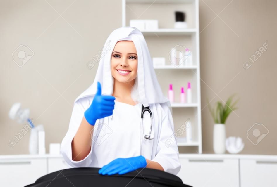Consulta em clínica de cosmetologia. Médico de beleza feminino em casaco branco e luvas pretas mostra o polegar para cima. Conceito de saúde.