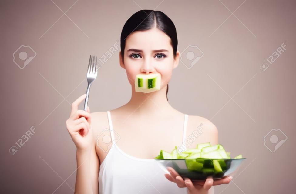 Una joven triste y hambrienta con una blusa blanca, sostiene en su mano un tenedor con una rodaja de pepino y un tazón con ensalada verde fresca, su boca está sellada con una cinta con la palabra comida, está perdiendo peso.