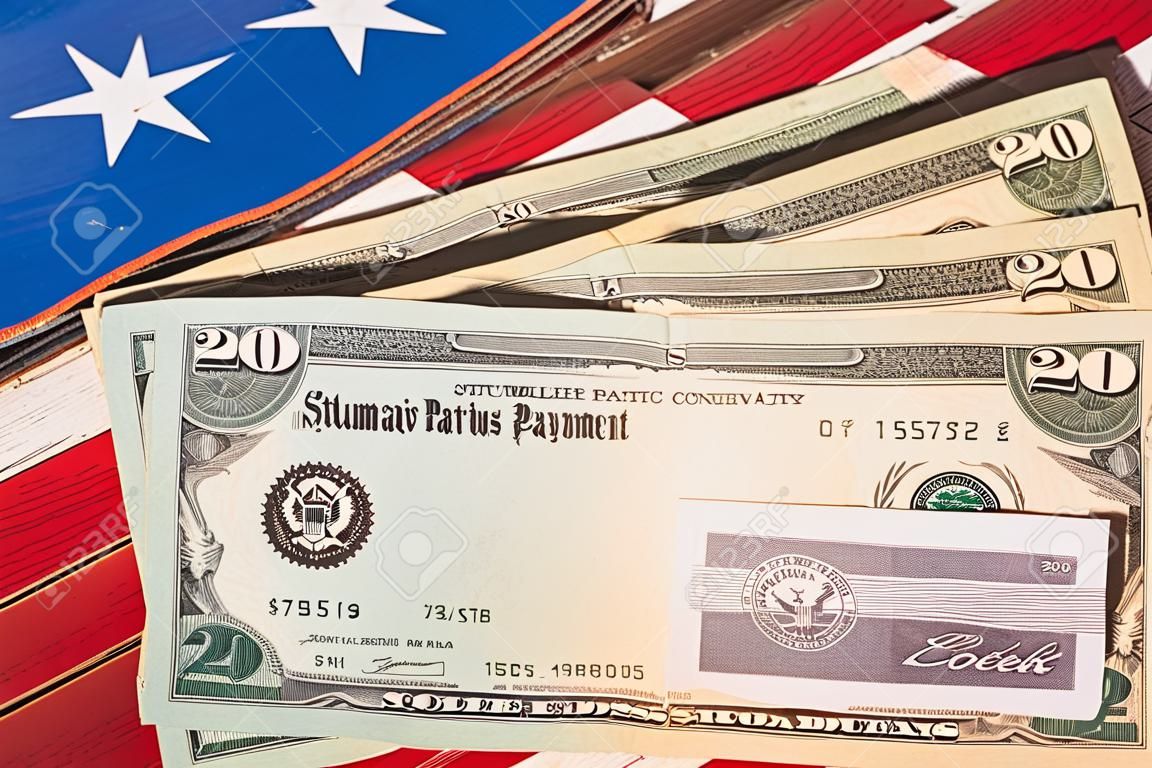 Pila de billetes de 20 dólares con cheque ilustrativo de pago de estímulo de coronavirus en la bandera de EE. UU.