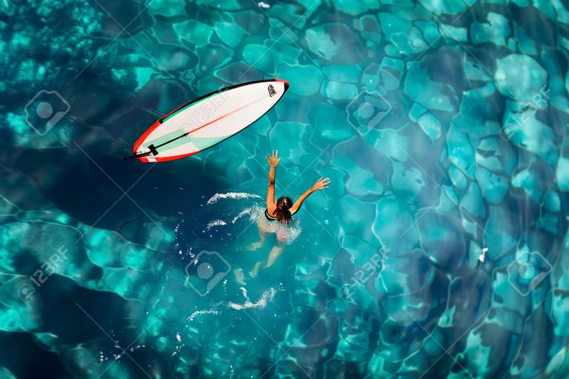Surf girl swim with surfboard in transparent ocean. Aerial top view