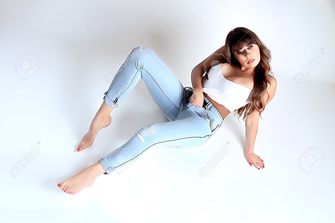 Portrait of attractive young woman with jeans trousers and white top. Well-built female model sitting on the ground.