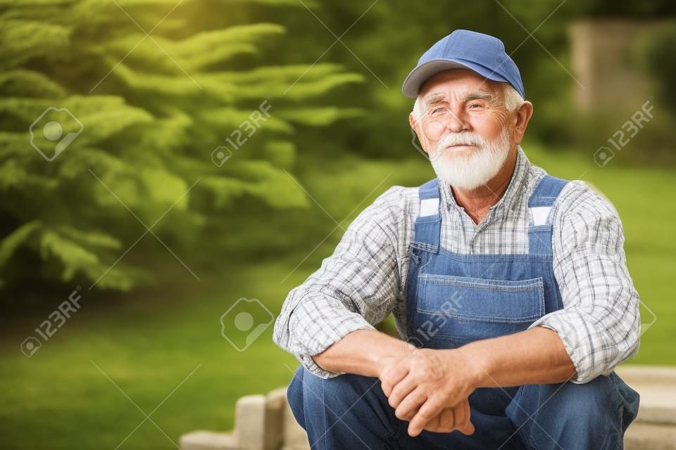 Senior gardener sitting on the steps in garden. Aged worker in overall and baseball cap resting after work. Portrait of old farmer