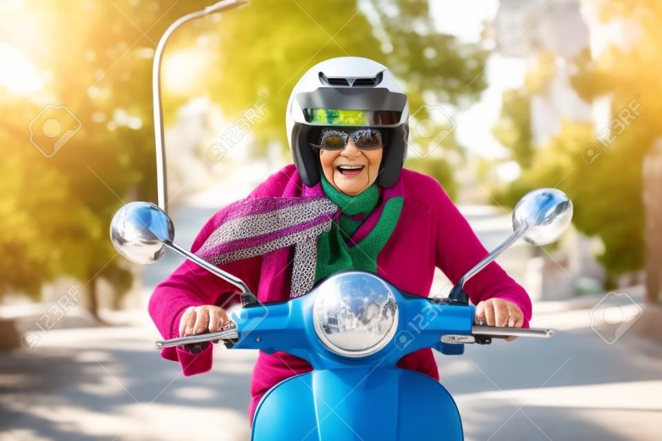 ヘルメット、サングラス、喜びの満面の笑みでカメラに直接彼女のスクーターに乗ってスカーフを身に着けてシニアの女性
