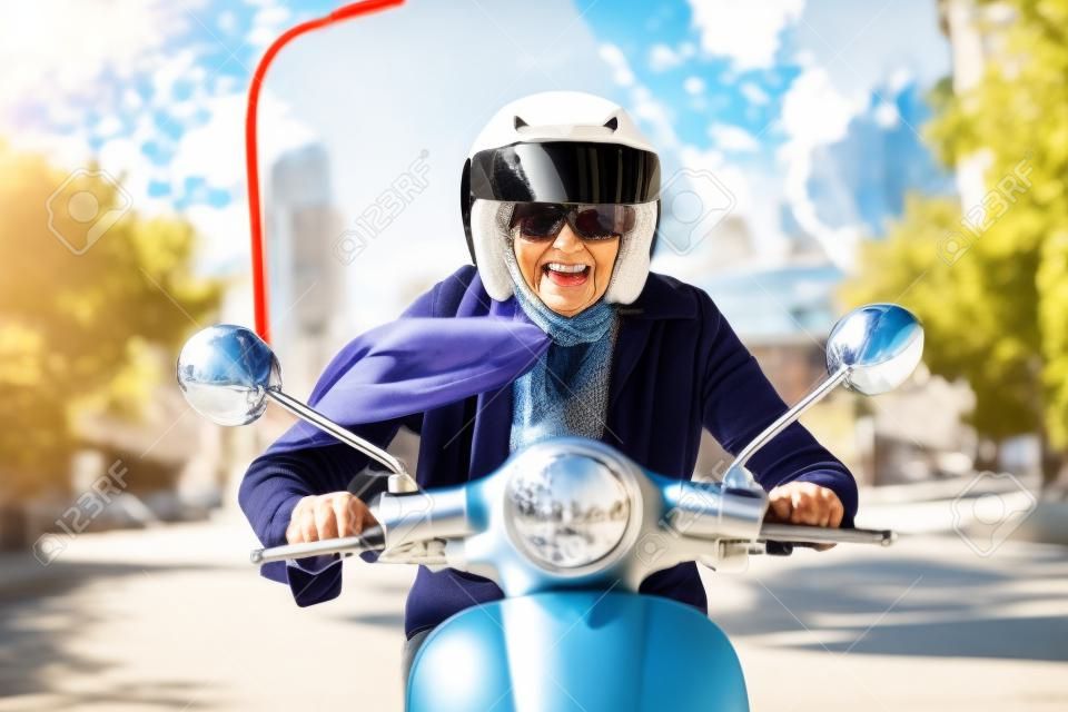 Senior lady wearing a helmet, sunglasses and a scarf riding her scooter directly at the camera with a wide smile of pleasure