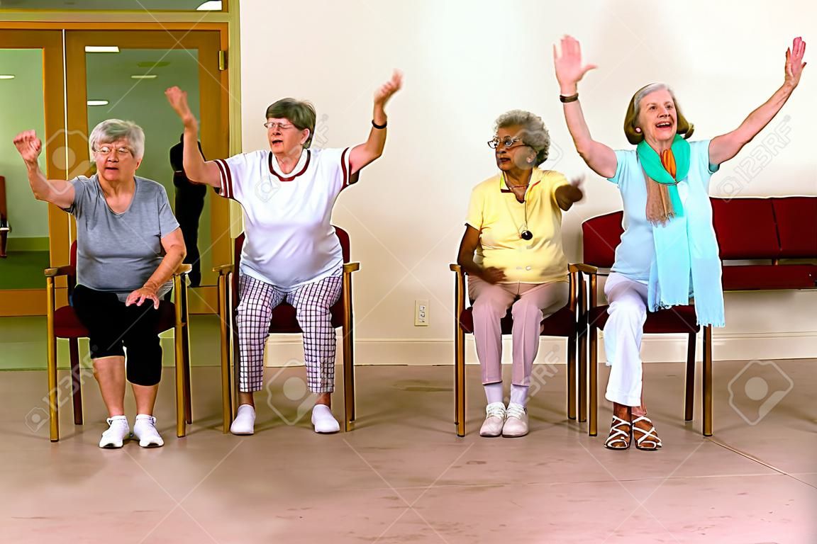 Group of four cheering senior women practicing light aerobic exercises with chairs for fitness class indoors