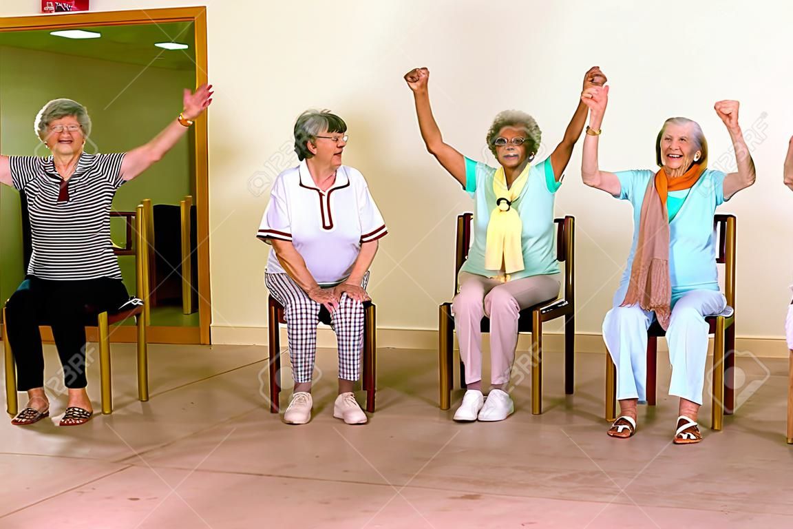 Group of four cheering senior women practicing light aerobic exercises with chairs for fitness class indoors