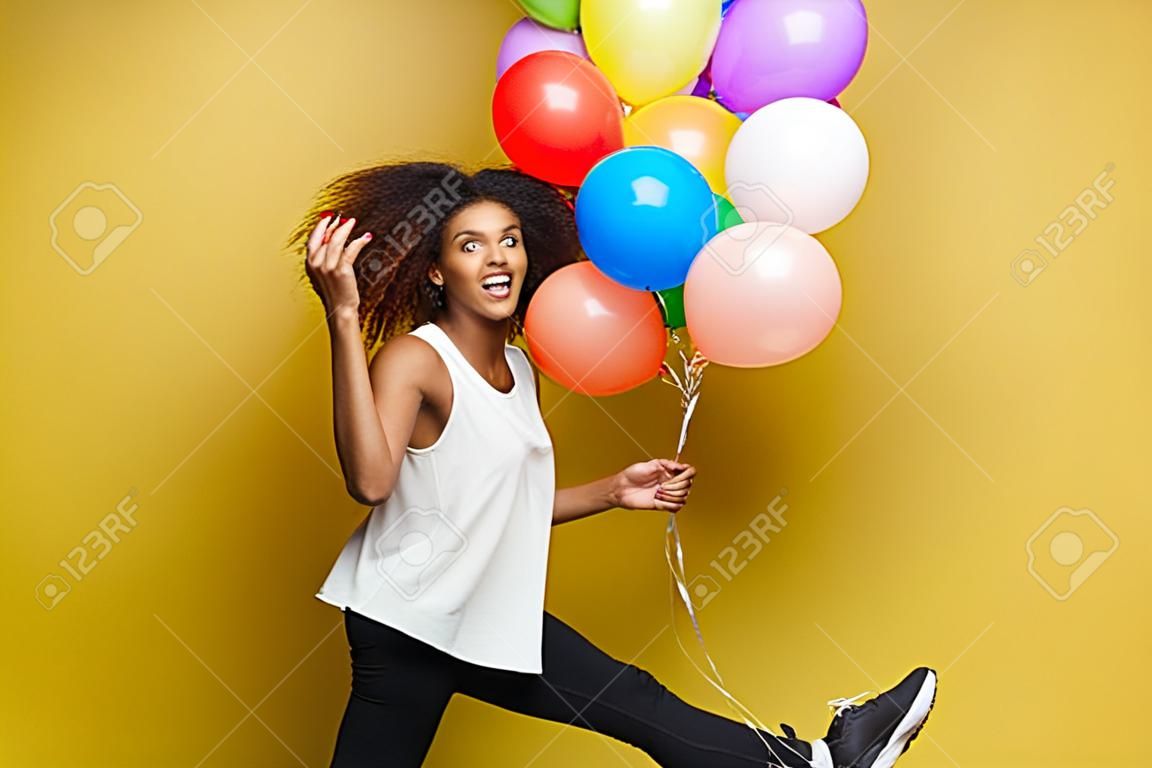 Concept de célébration - Close up Portrait heureux jeune belle femme africaine avec un t-shirt blanc en cours d'exécution avec un ballon de fête coloré. Fond de studio Pastel jaune