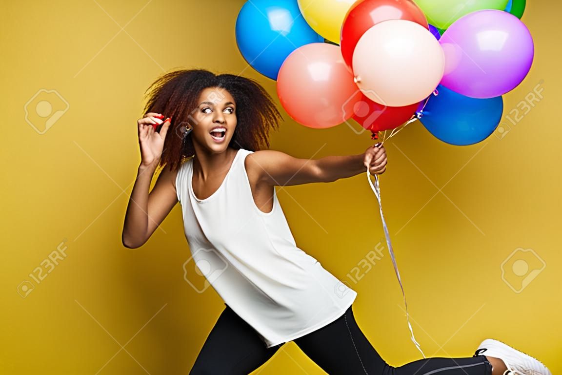 Feier-Konzept - nahes hohes glückliche junge schöne afrikanische Frau des Porträts mit dem weißen T-Shirt, das mit buntem Parteiballon läuft. Gelber Pastellstudio Hintergrund