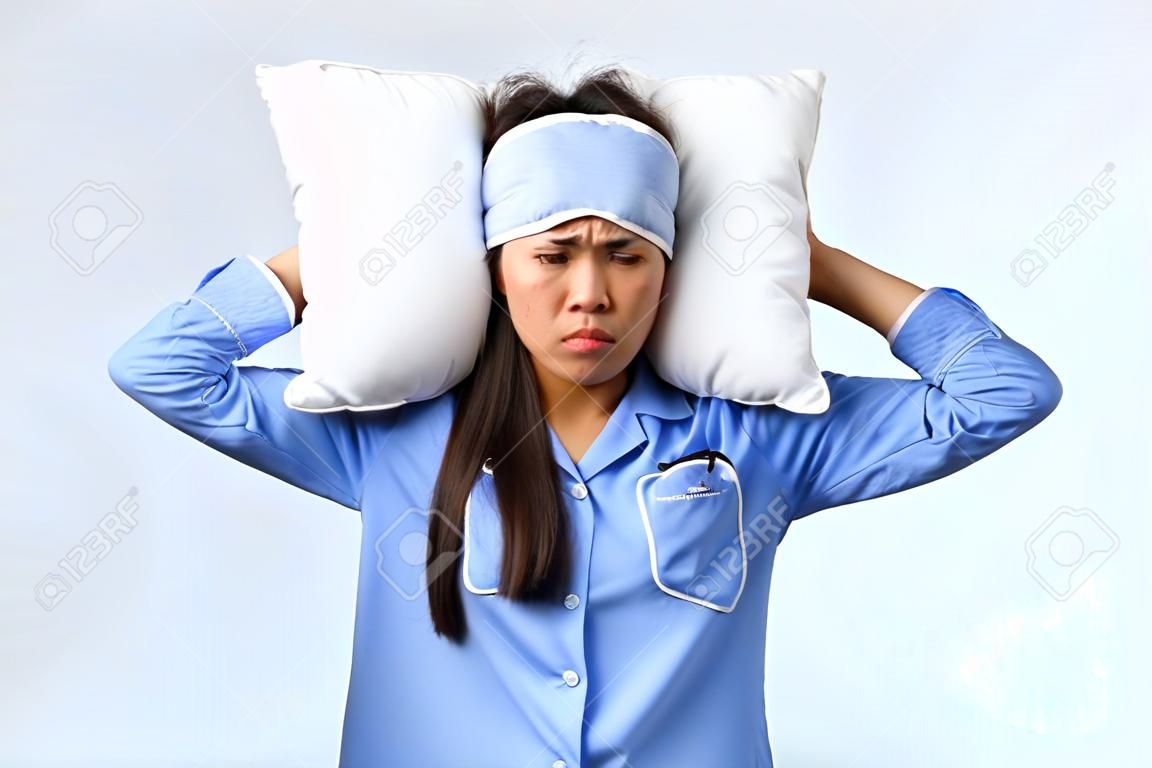 Annoyed and mad grumpy asian girl with insomnia, lying in bed in pajamas and sleeping mask, cover ears with pillow and look bothered, irritated with friend snoring loud at night, white background