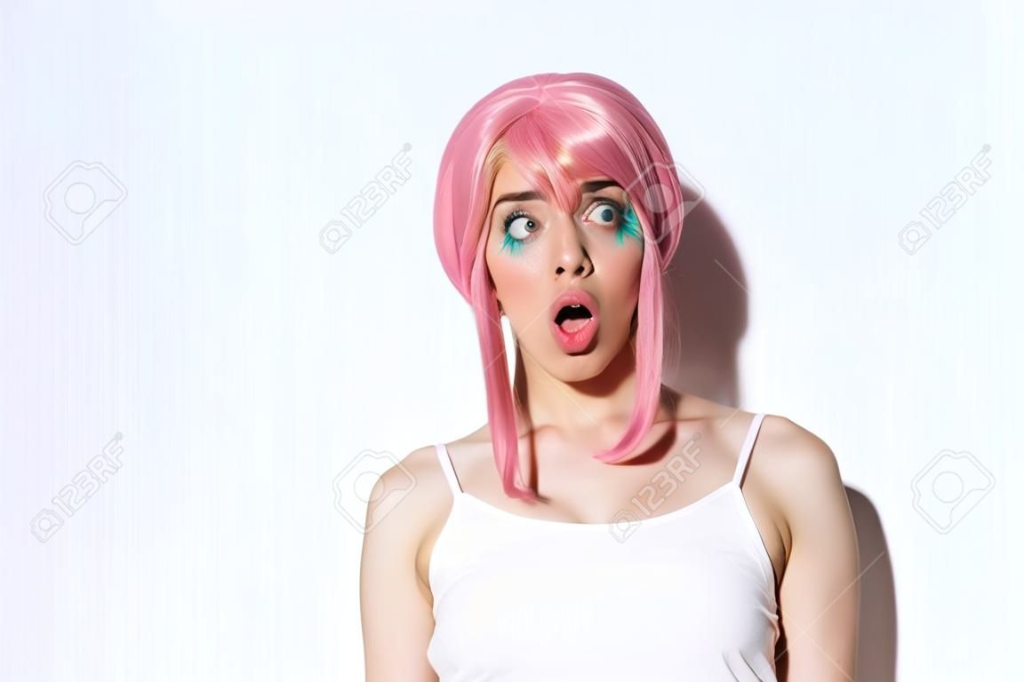 Gros plan d'une jolie fille impressionnée en perruque rose, bouche ouverte avec admiration et regardant à gauche, debout sur fond blanc