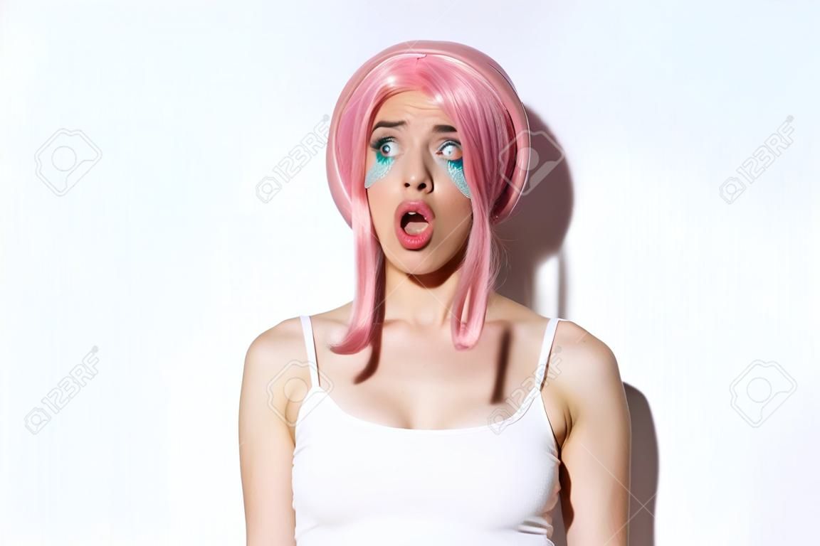 Gros plan d'une jolie fille impressionnée en perruque rose, bouche ouverte avec admiration et regardant à gauche, debout sur fond blanc