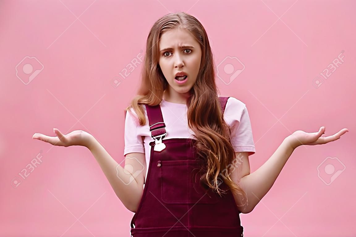 So what, no big deal. Rude and impolite arrogant young girl with high ego in overalls spreading hands sideways in questioned, bothered gesture expressing confusion, feeling pissed with stupid question