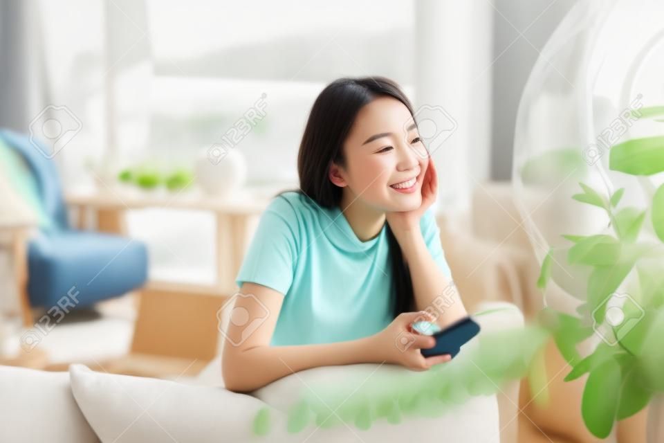 Marzycielska azjatka spędza czas w domu, trzymając smartfon i siedząc na kanapie, uśmiechając się patrząc w okno