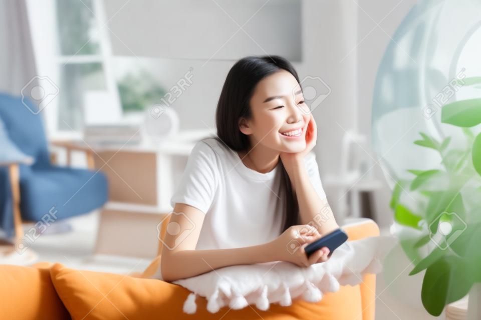 꿈꾸는 아시아 소녀는 집에서 시간을 보내고, 스마트폰을 들고 소파에 앉아 창문을 보며 웃고 있습니다.
