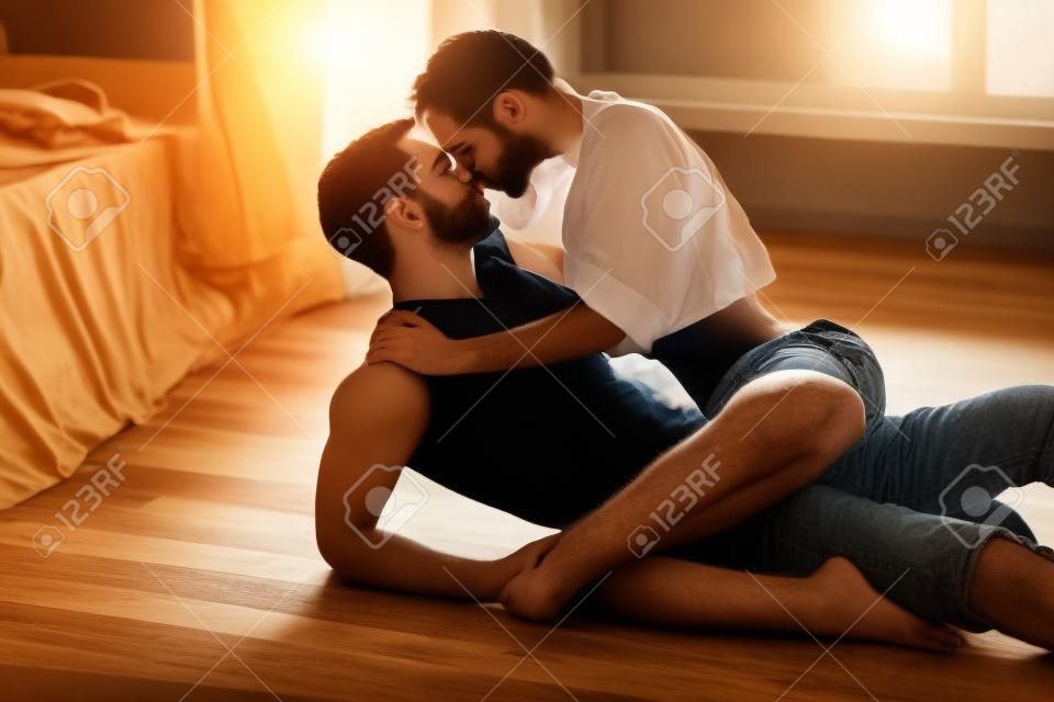 Leidenschaftliche Paar küsst, Jungen und Mädchen sitzen auf Holzboden in der Nähe des zerwühlten Bett gegenüber dem Fenster.