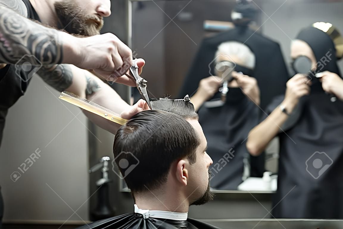 Unmatched barber with a big beard is cutting the hair ends of his client in the black cutting hair cape in the barbershop. Customer has hairgrips on the head. The both blurry reflected in the mirror.