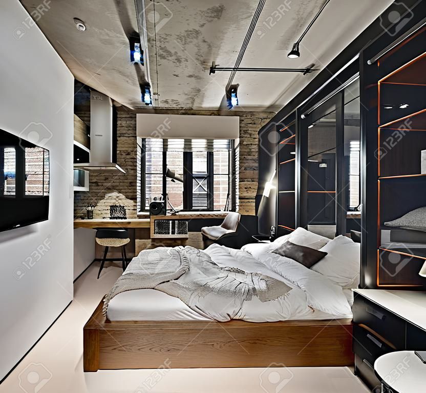 Bedroom in a loft style with brick wall and concrete ceiling. There is a TV, bed with pillows, lamps with lampshades, wardrobe with glass sliding doors, tables, armchair, parquet with a carpet.