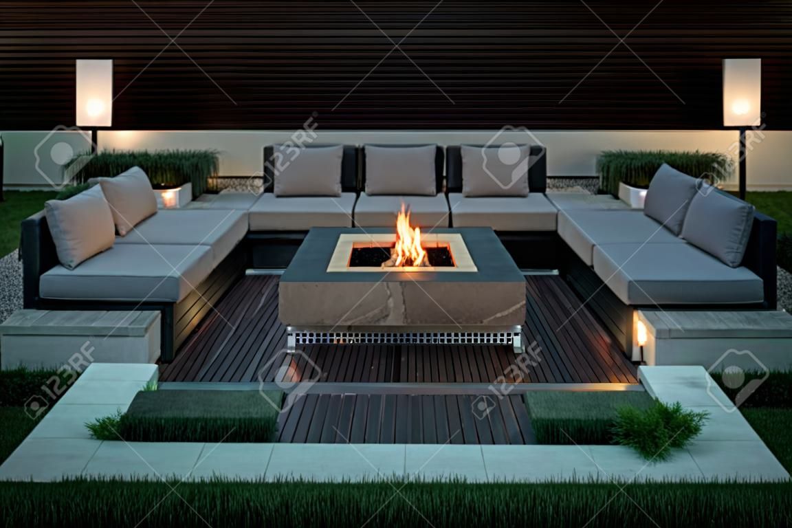 Zone for relax with a wooden floor and a tiled stair outdoors. There is a burning fire pit, gray sofas and armchairs, plaid, luminous lamps. Horizontal.