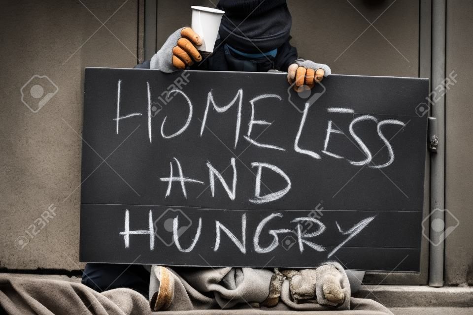 Tramp sitting on the street with a cup for money and holding a sign with text Homeless and hungry