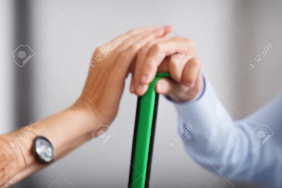 ぼやけた背景に対して歩行棒を持つ先輩の手を握る介護者のクローズアップ