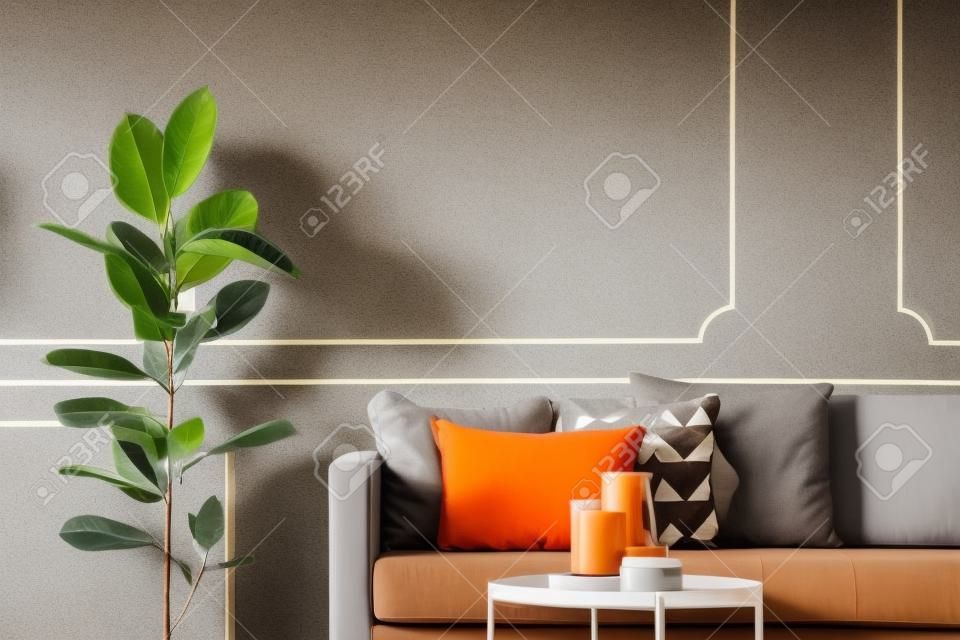 Ficus accanto al divano marrone con cuscini arancioni all'interno del soggiorno grigio con tavolo. Foto reale