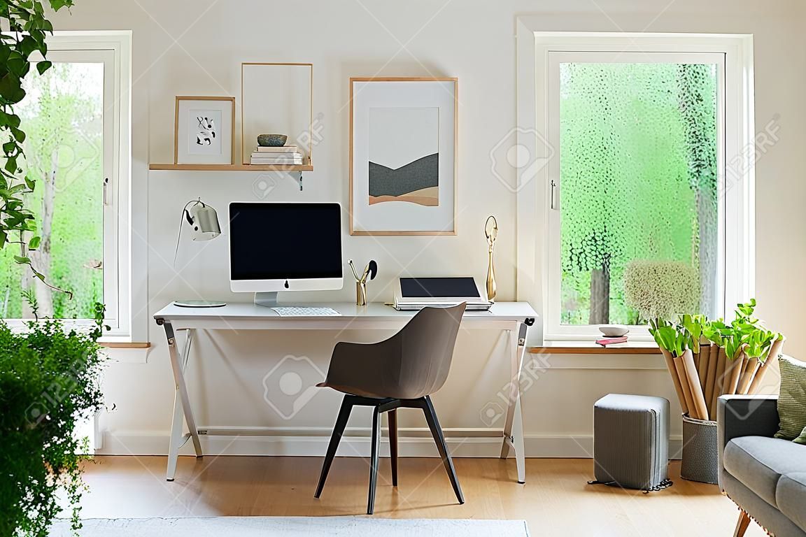 Grauer Stuhl am Schreibtisch mit Desktop-Computer in scandi offenem Rauminnenraum mit Fenstern. Echtes Foto