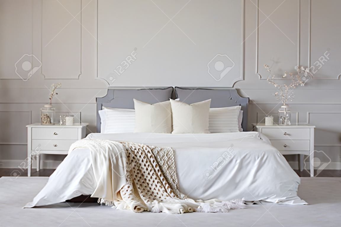 Łóżko małżeńskie z białą pościelą i kocem pomiędzy dwoma drewnianymi stolikami nocnymi kwiaty w wazonach