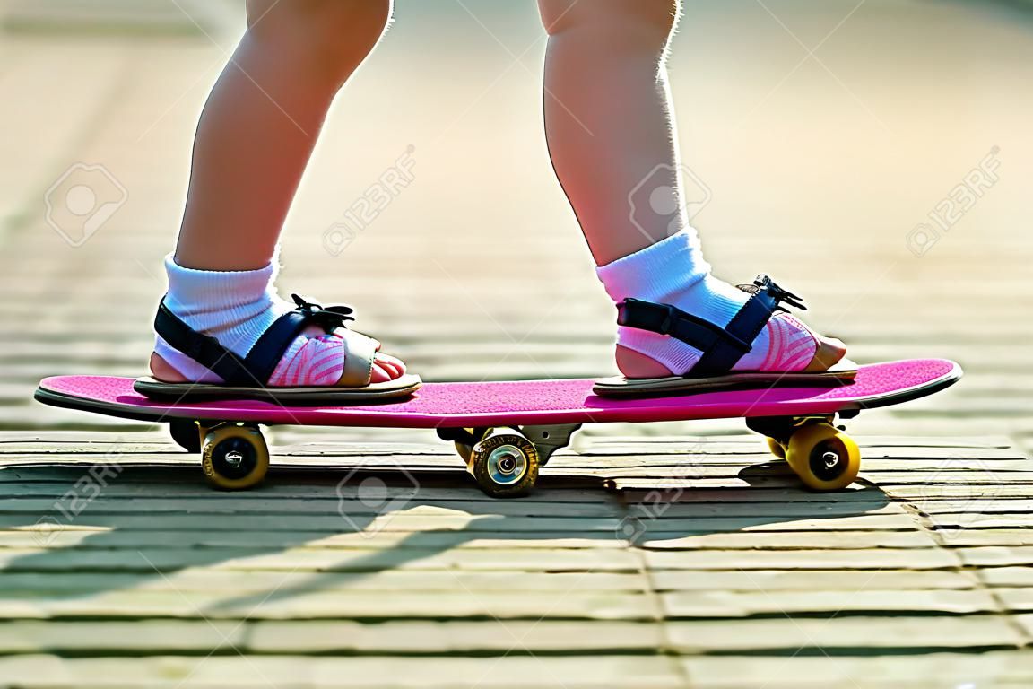 Schlanke Beine des Kindes in den weißen Socken und in den schwarzen Sandalen auf dem rosa Skateboard des Plastiks auf hellem sonnigem Sommer verschwommener Kopierraumpflasterhintergrund. Outdoor-Aktivitäten und gesundes Lifestyle-Konzept.