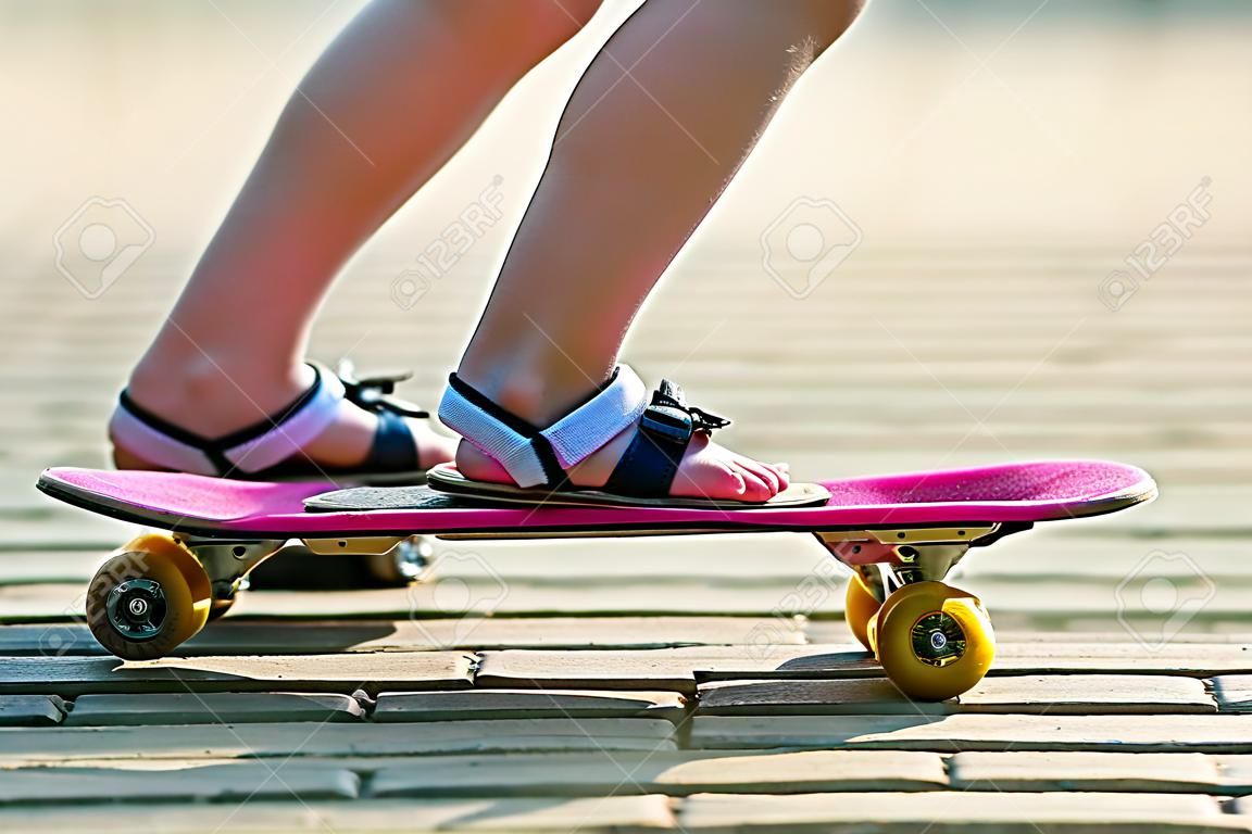 在白色袜子的儿童亭亭玉立的腿和在塑料桃红色滑板的黑凉鞋在明亮的晴朗的夏天弄脏了拷贝空间路面背景。户外活动和健康的生活方式的概念。
