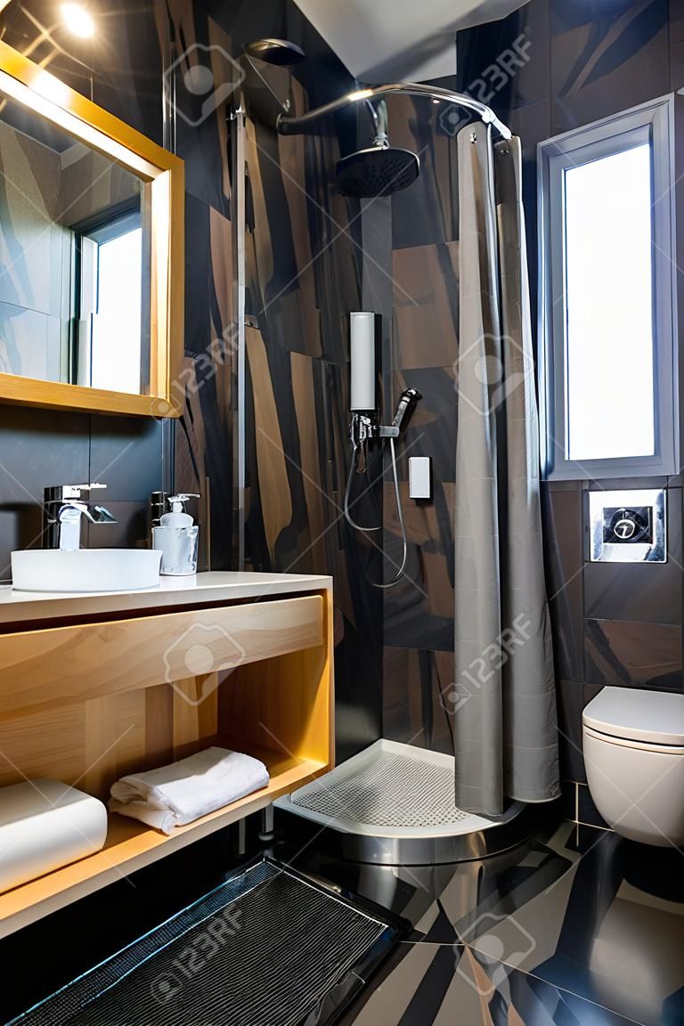 Interior do moderno banheiro elegante com paredes de azulejos pretos, lugar de chuveiro cortina e móveis de madeira com lavatório e grande espelho iluminado.