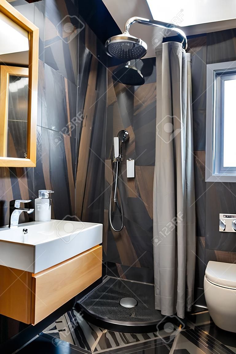 Interior do moderno banheiro elegante com paredes de azulejos pretos, lugar de chuveiro cortina e móveis de madeira com lavatório e grande espelho iluminado.