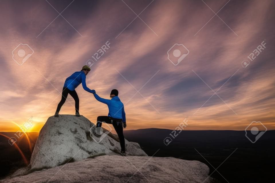 Man and woman hikers helping each other to climb stone at sunset in mountains. Couple climbing on high rock in evening nature. Tourism, traveling and healthy lifestyle concept.